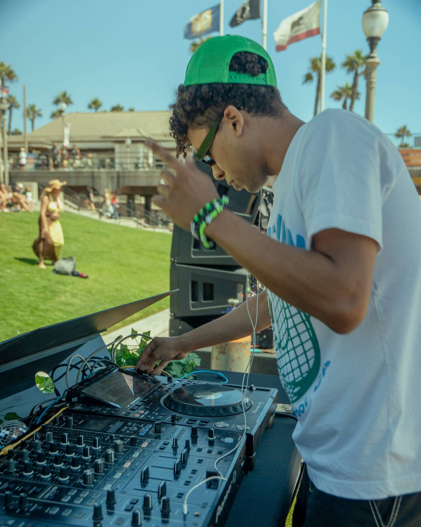 DJ in a green cap mixing at the decks with the Huntington Beach pier behind him
