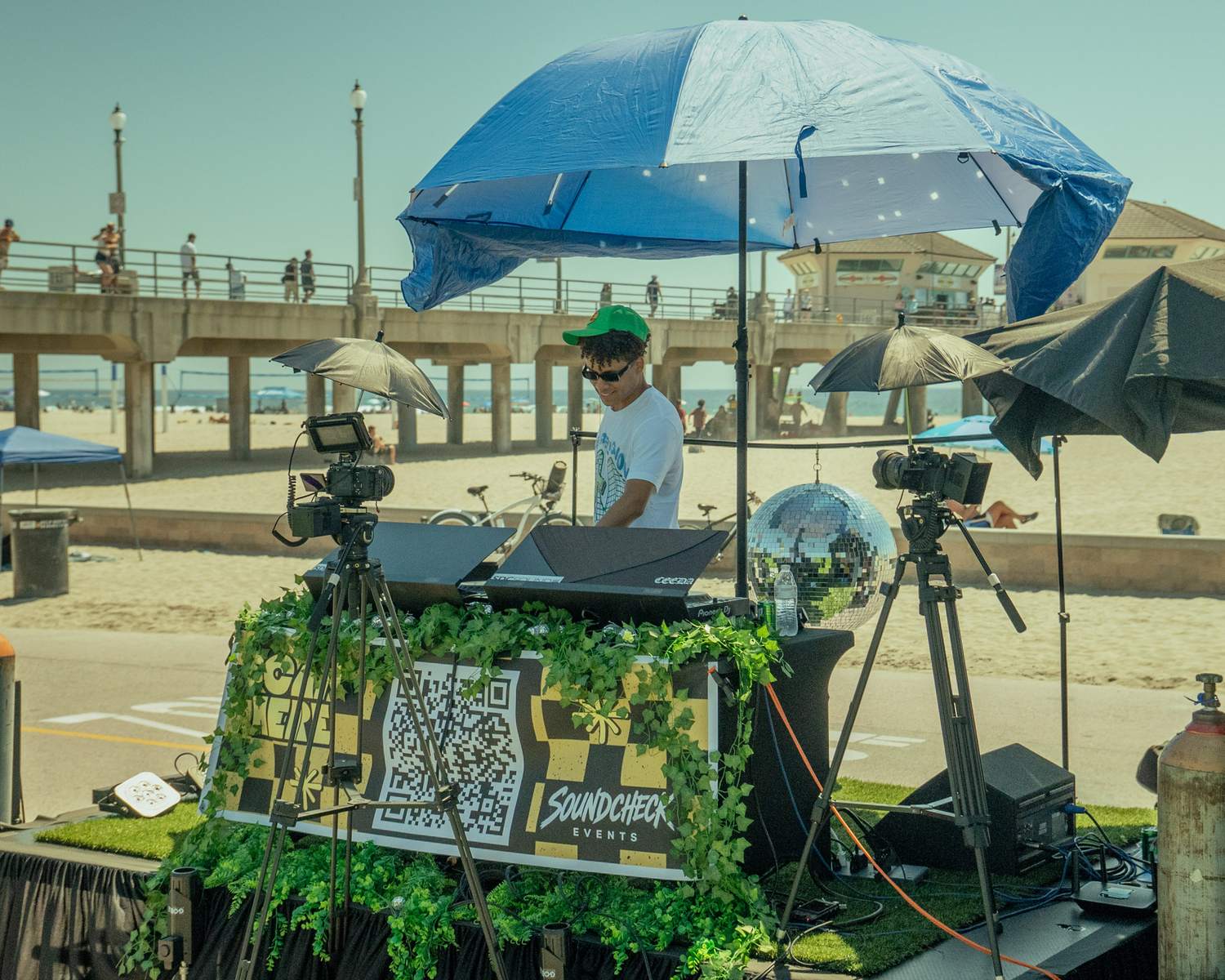 DJ booth wrapped in greenery with a Soundcheck banner on the beach under a blue umbrella