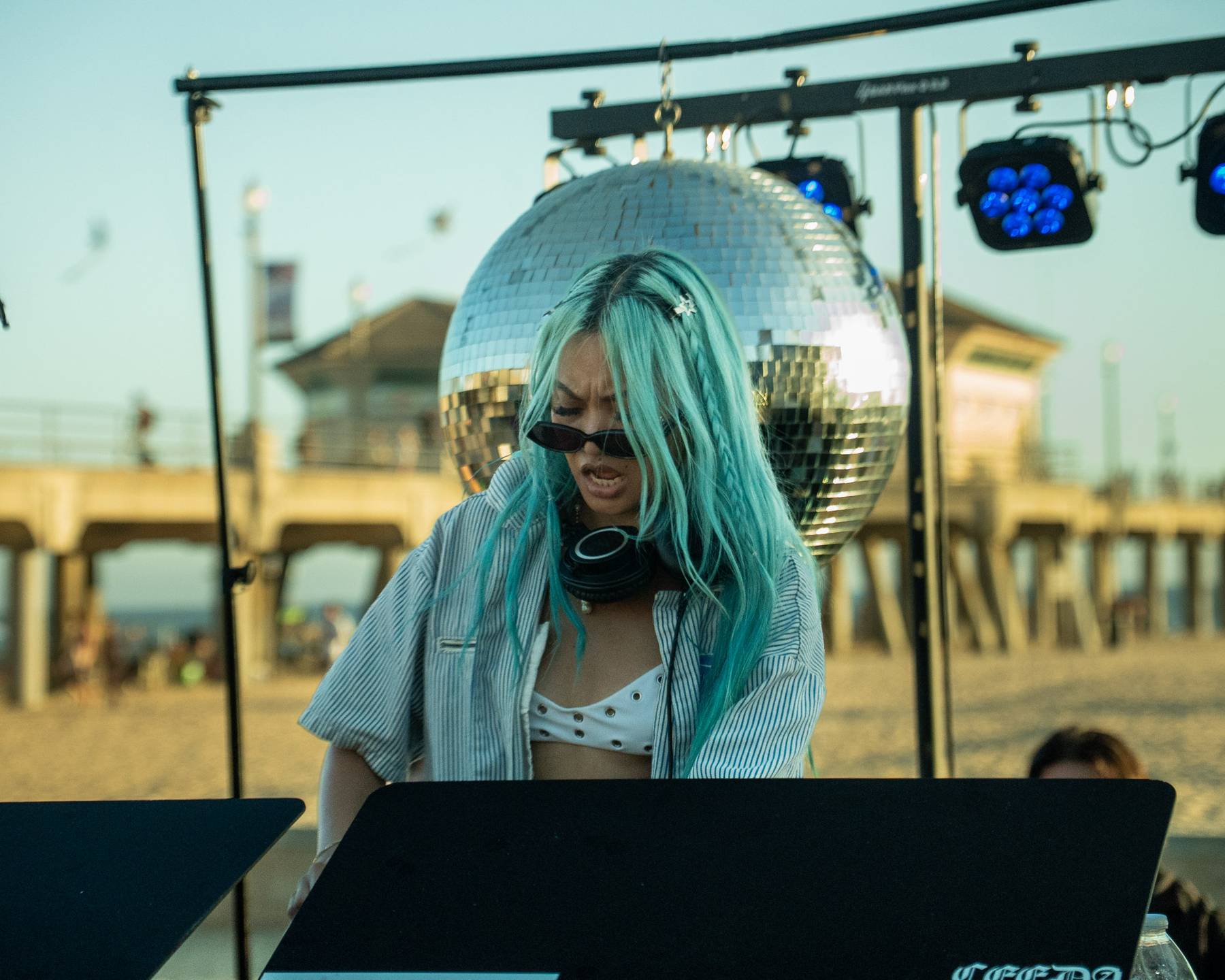 DJ with teal hair and a disco ball behind her at the pier