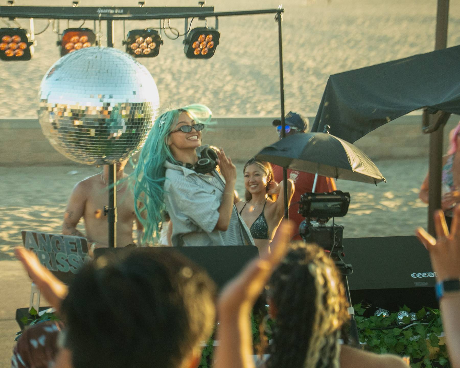 DJ with teal hair behind the decks as the crowd cheers, disco ball overhead