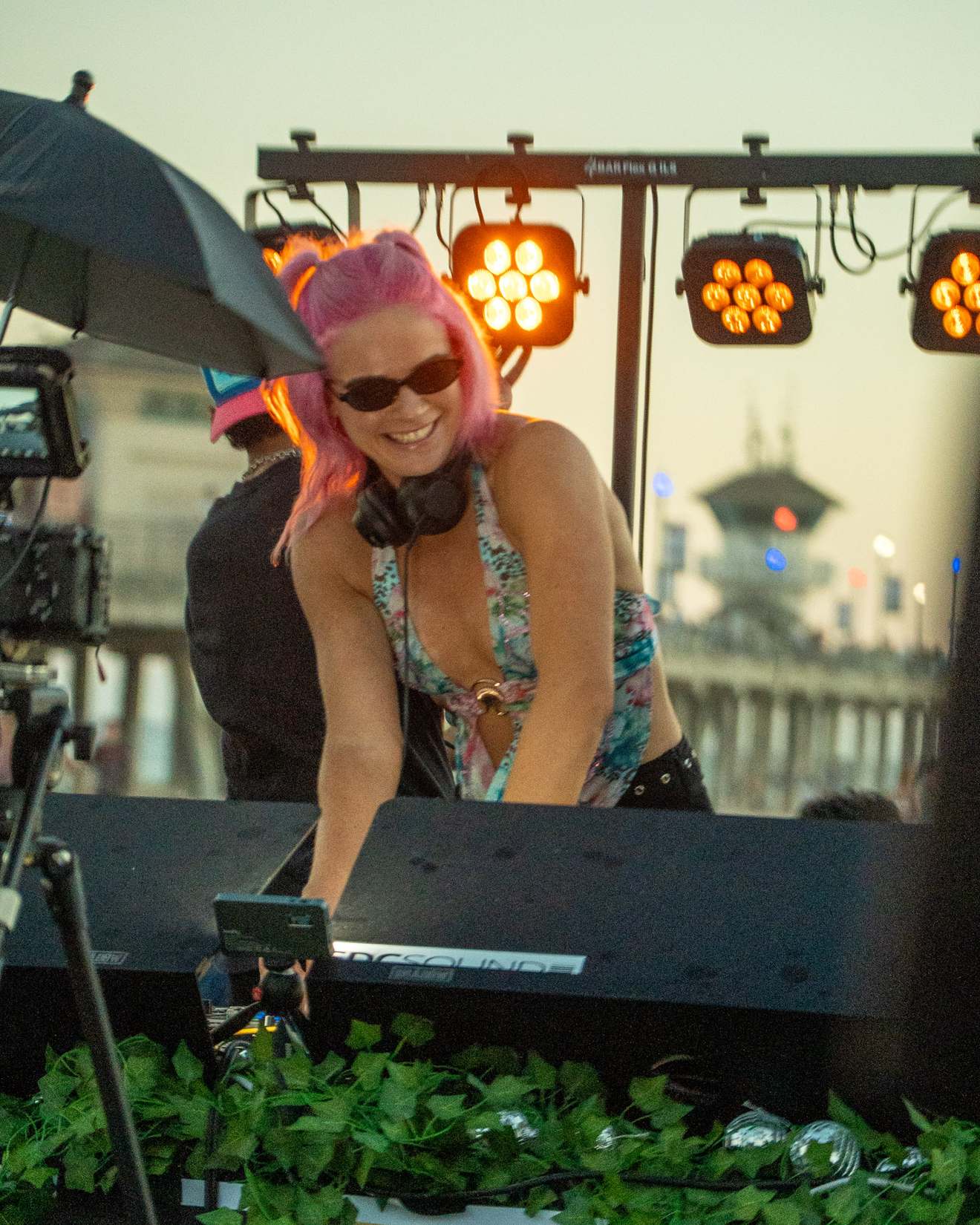 DJ with pink hair leaning over the decks under stage lights at dusk